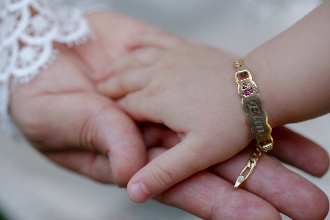 Close-up of a baby holding an adults hand, featuring a gold bracelet with engraved name.